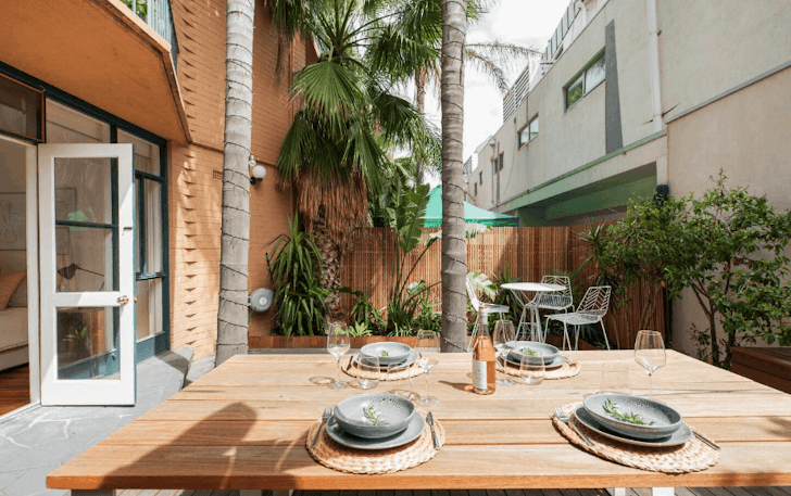 A light timber table sits outside in an Airbnb Melbourne courtyard, plated with tableware and cutlery.