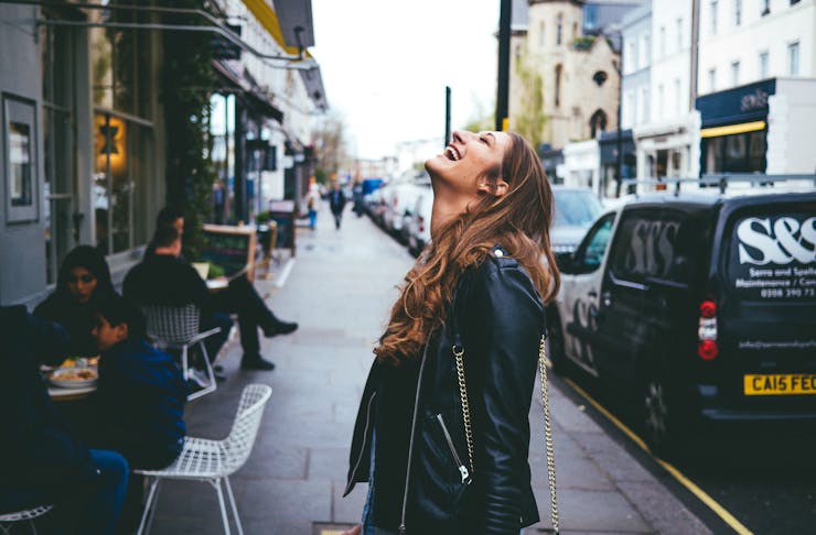 Young woman in a leather jacket on a busy street smiling and looking up at the sky.