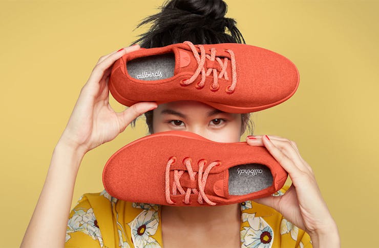 A girl holds up a red pair or Allbirds in front of a yellow background