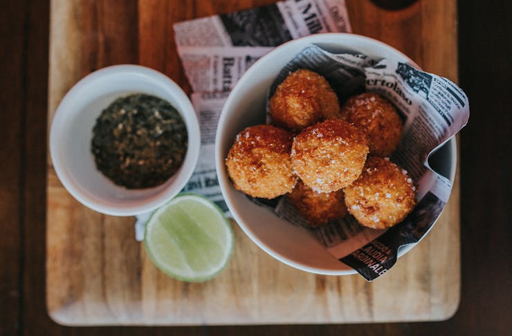 A bowl of arancini bowls sits on a wooden board with a small bowl of sauce.