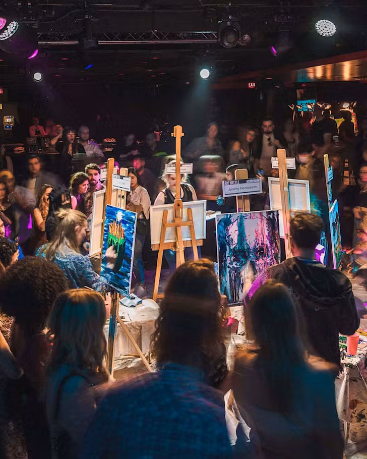 Artists standing at easle-propped canvases, battle it out under bright colourful lights at an Art Battle night.