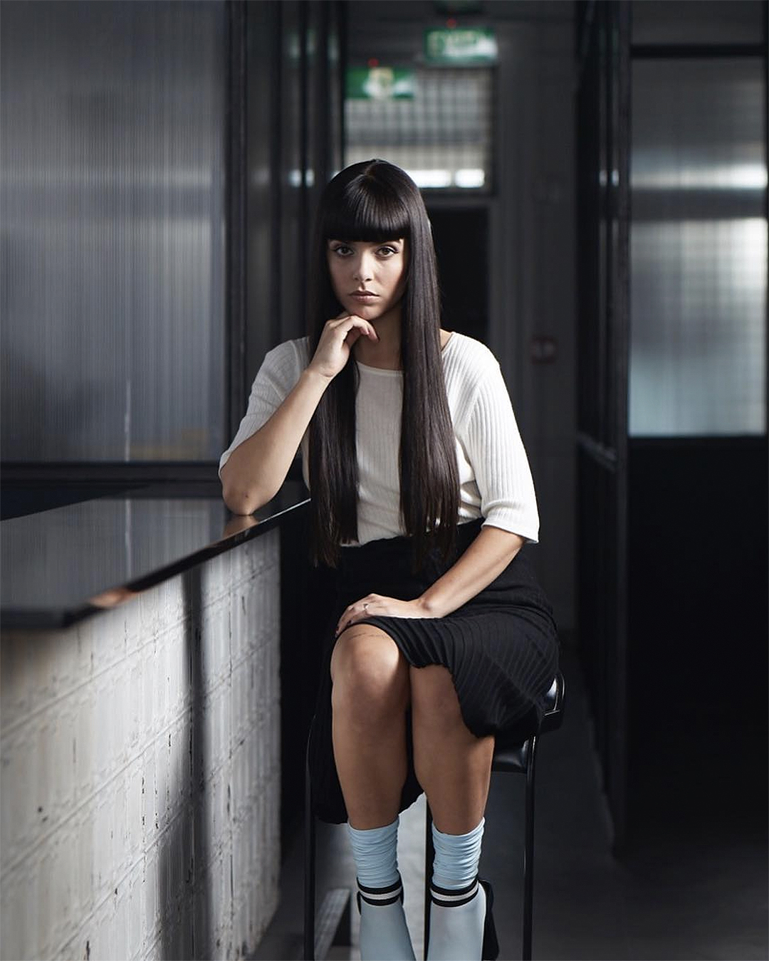 A woman sits on a stool with impeccably combed long dark hair.
