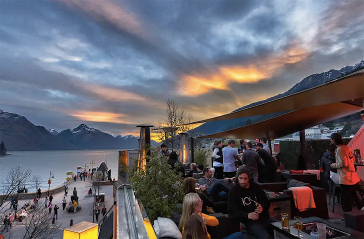 People frequent a beautiful rooftop bar in front of a stormy Queenstown sky.