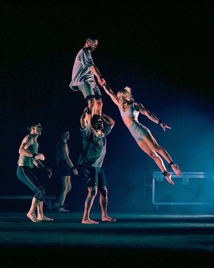A dancer is lifted mid-air on stage by another dancer on top of another dancer's shoulders. Two other dancers, entirely, watches on.