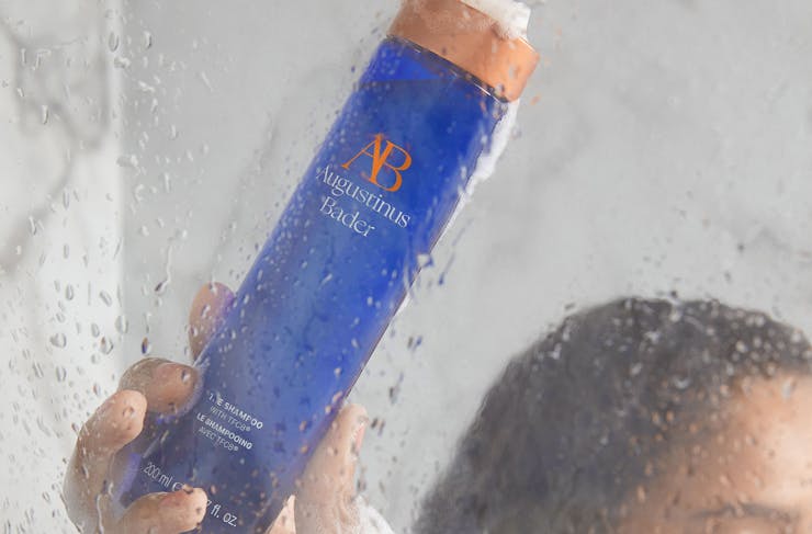 A person in the shower holding a bottle of Augustinus Bader shampoo. 