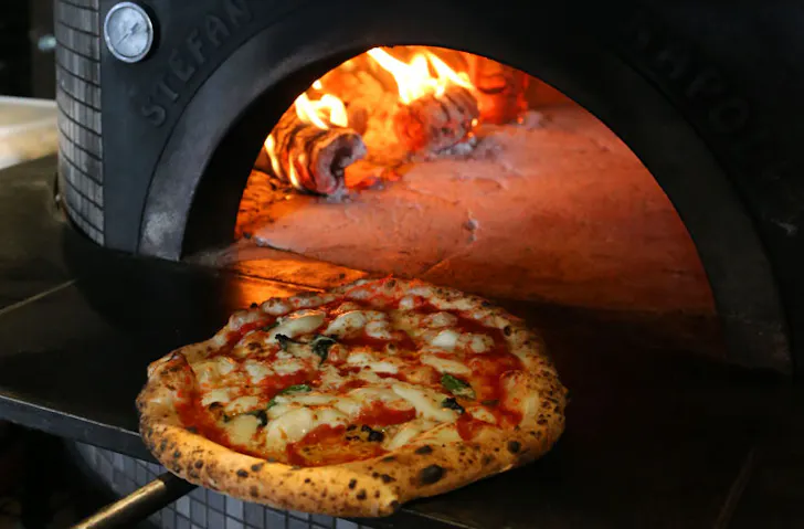 A Napoli-style Margherita pizza being retrieved from a stone pizza oven. 