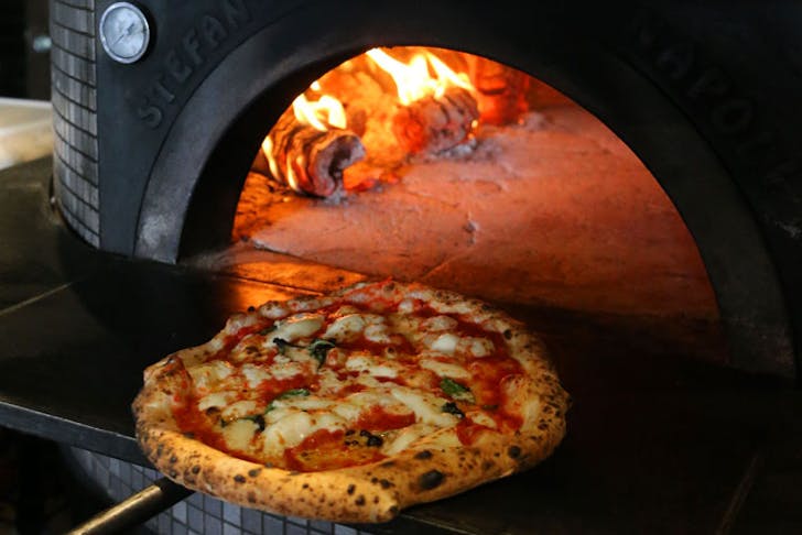 A Napoli-style Margherita pizza being retrieved from a stone pizza oven. 