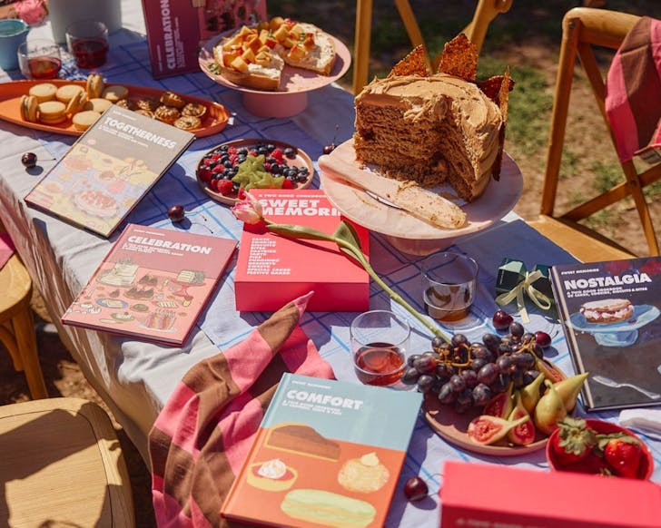 A mid shot image of an outdoor dining table aesthetically decorated with baked goods such as cakes and biscuits with Two Good Co.'s cookbooks laid out amongst the table. The light is shining nicely onto the table and it is decorated with fruit and a nice tablecloth.