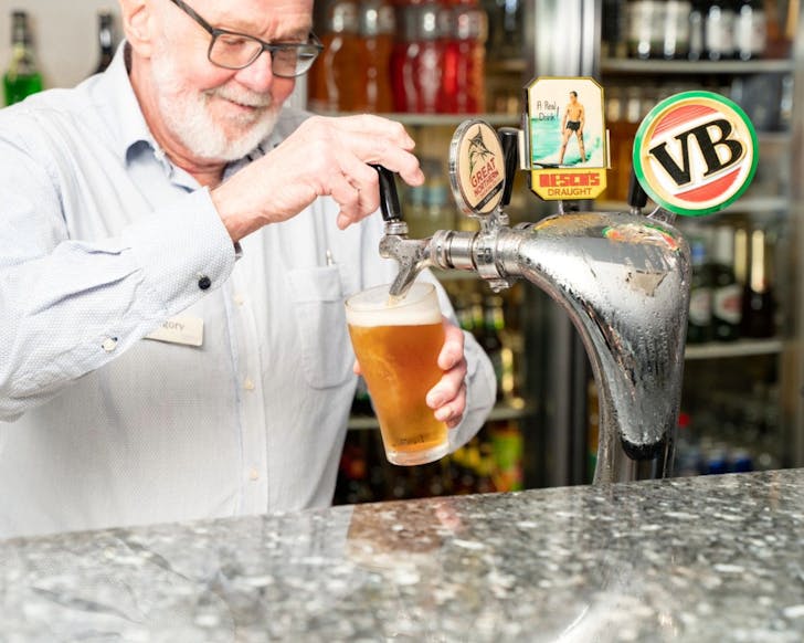An older man is smiling while filling up his glass of beer from the VB tap at the bar. 