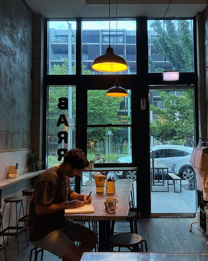 A wide image of the interior of Bario Collective Coffee where the design is minimalistic and there is a large window wall in the background of the image leading out to the street. A single man sits down on the cafe benches, invested in his journal writing.