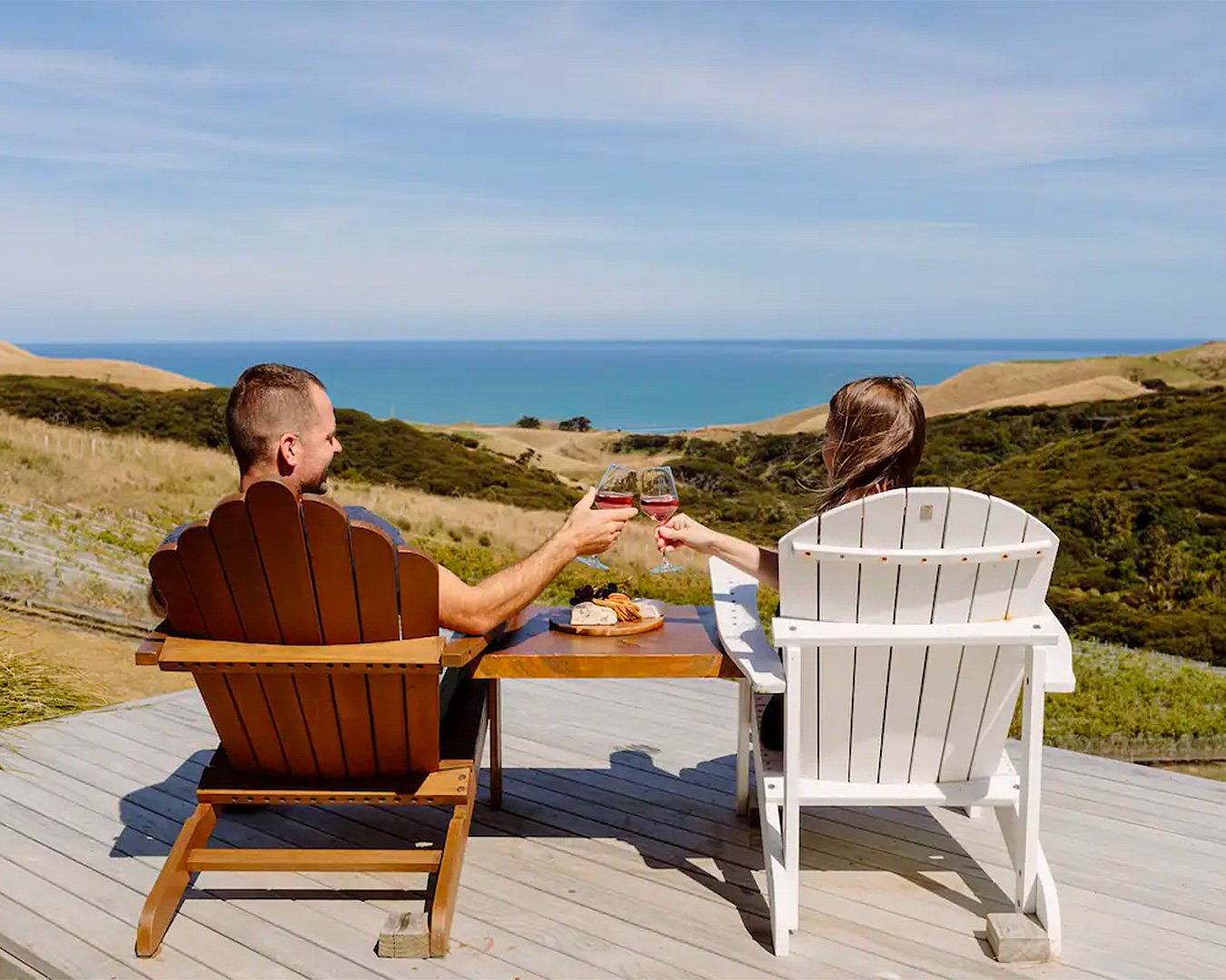 Two people enjoy wine on a deck overlook an exquisite beach view at one of the most romantic getaways near Auckland. 