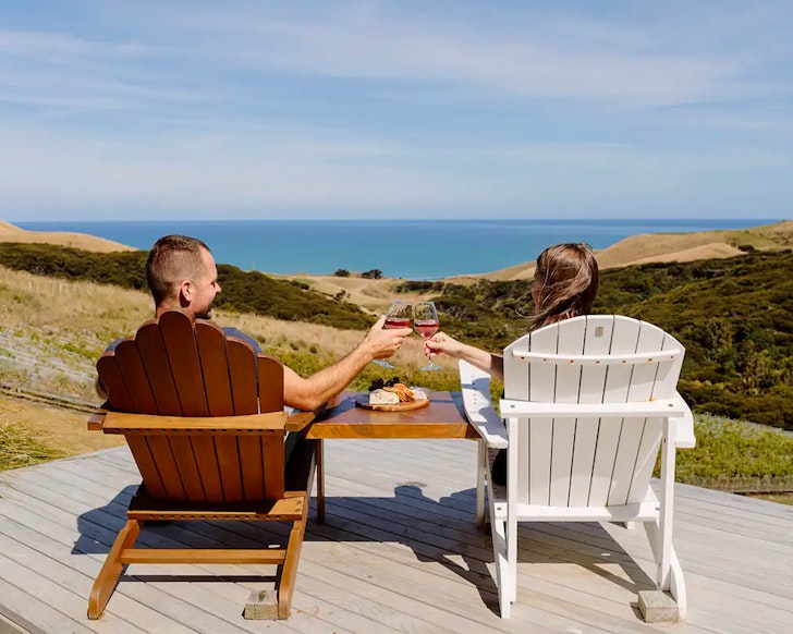 A couple clinks wine glasses overlooking a view at Barrique Studio @ Barrelled Wines Raglan one of the best romantic stays near Auckland