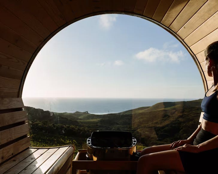 Someone looks out toward the west coast of the Waikato, while sitting in a glass-fronted barrel sauna.