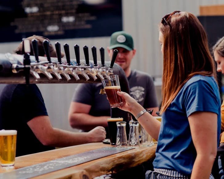 A mid shot of a lady filling up a glass of beer on tap. Behind her, and slightly blurred out of focus, are other people standing around the beer tap and drinking.