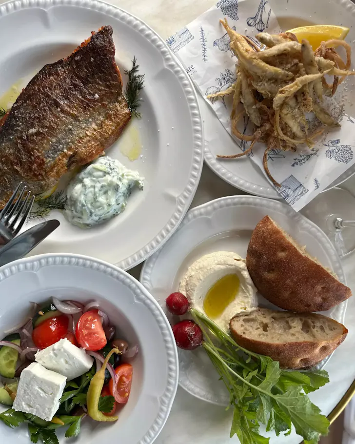 Overhead spread of grilled fish with herb sauce, fried whitebait, Greek salad with feta, and fresh bread with whipped dip served on white plates.
