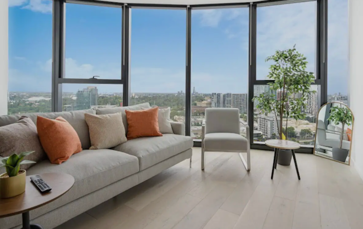 A large grey two seater sofa sits in a Airbnb Melbourne living room with floor to ceiling windows overlooking the surrounding landscape.