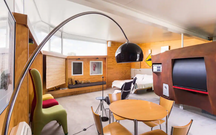 This Airbnb Melbourne living room is almost a circle, with timber walls enclosing the space. A large TV screen sits in an arched TV unit, with three sofa chairs sitting opposite it.