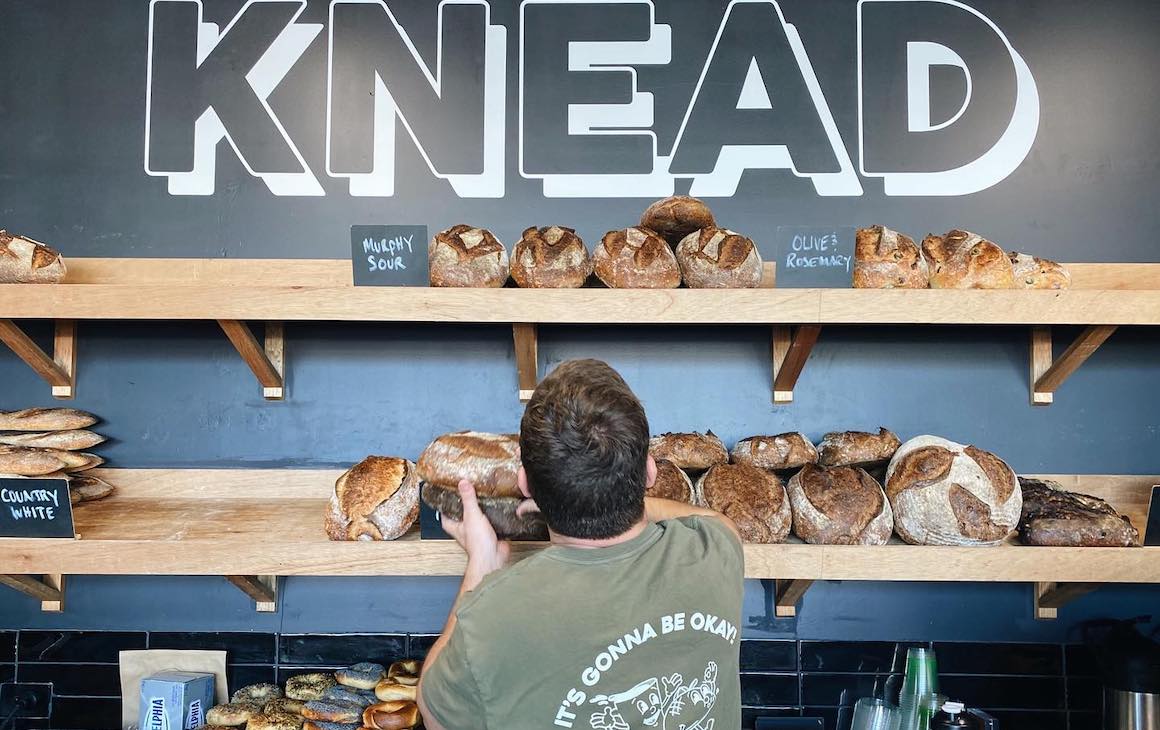 someone picking fresh loaves from the shelf with KNEAD written on the wall above