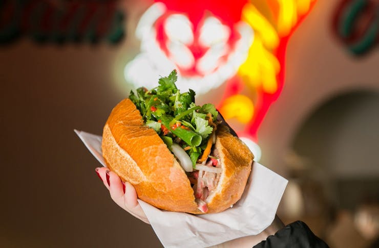 A person holding a banh mi roll, with a red and yellow neon light in the background. 