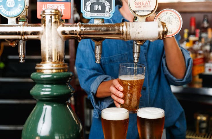 A bar person in a blue denim shirt pours schooners of beer from beer taps. 