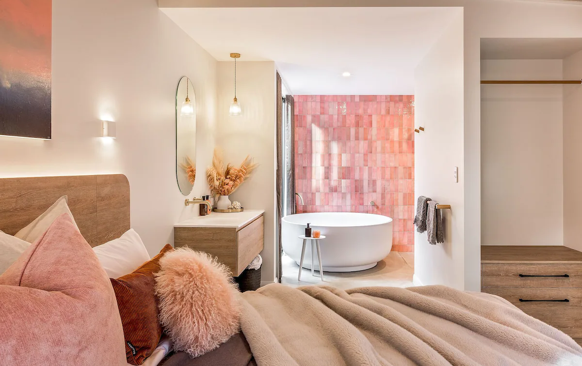 overlooking a bed piled high with cushions is a bathroom with a freestanding tub and a pink tile feature wall