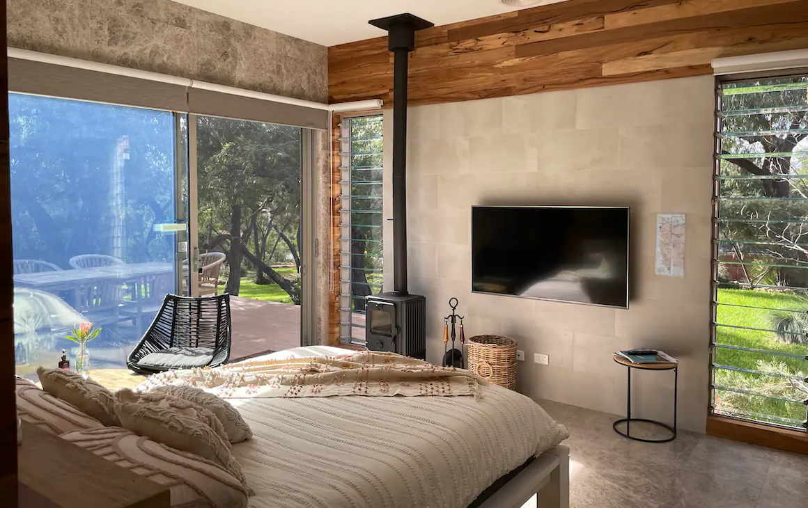 bed faces a wall with a rustic fireplace and a tv. The windows overlook bushland. 