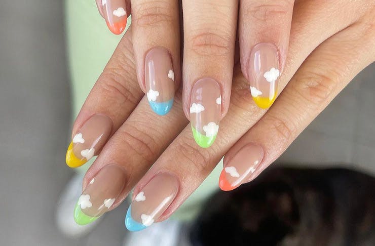 Hands showing nail art featuring blue, green, and yellow tips with soft clouds. 