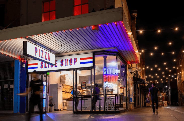 A neon-lit pizza shop on a corner in the evening. A sign reads Slice Shop, considered some of the best pizza in Melbourne. 