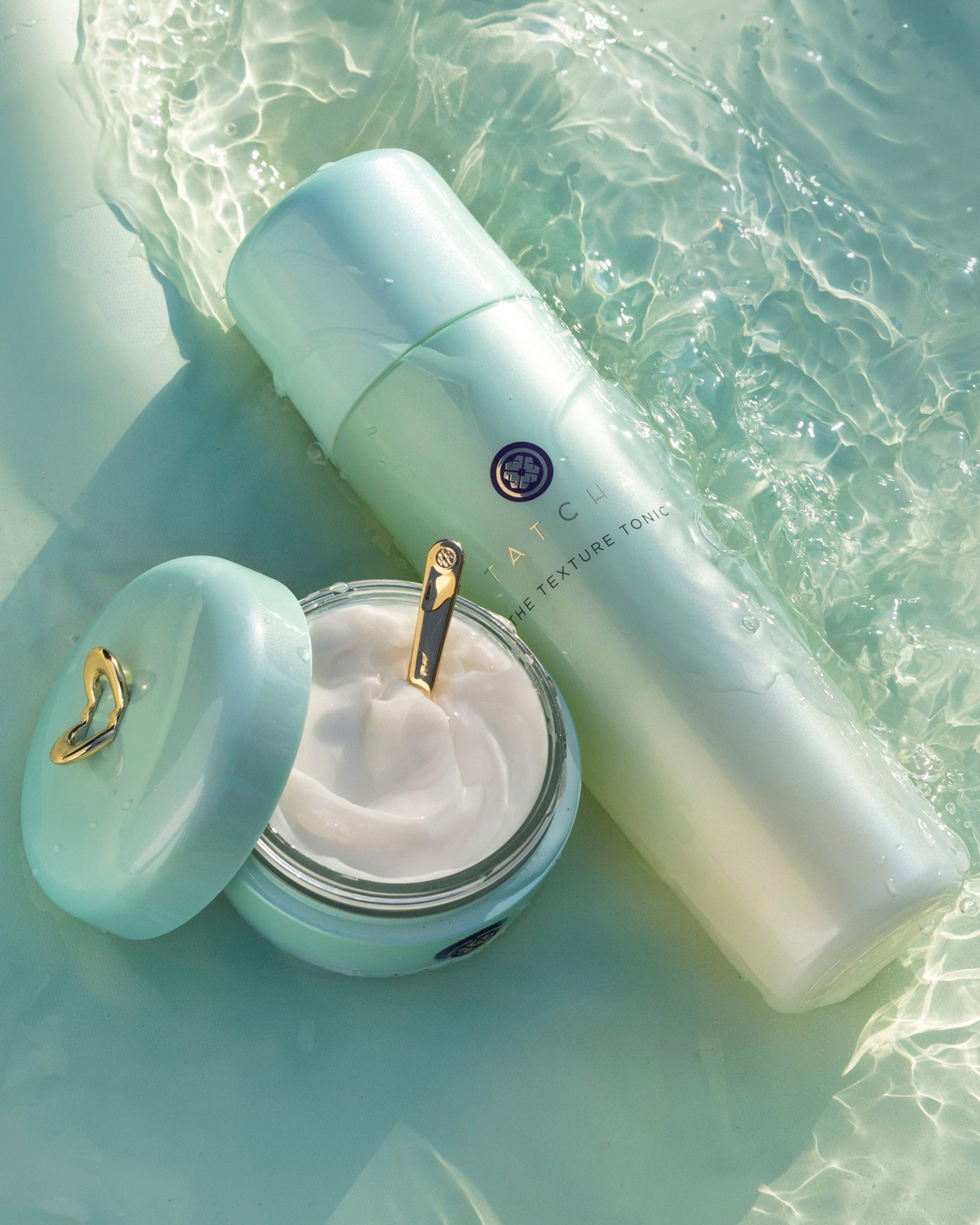 Tatcha bottles are seen in water with the lid off and a golden spoon protruding.