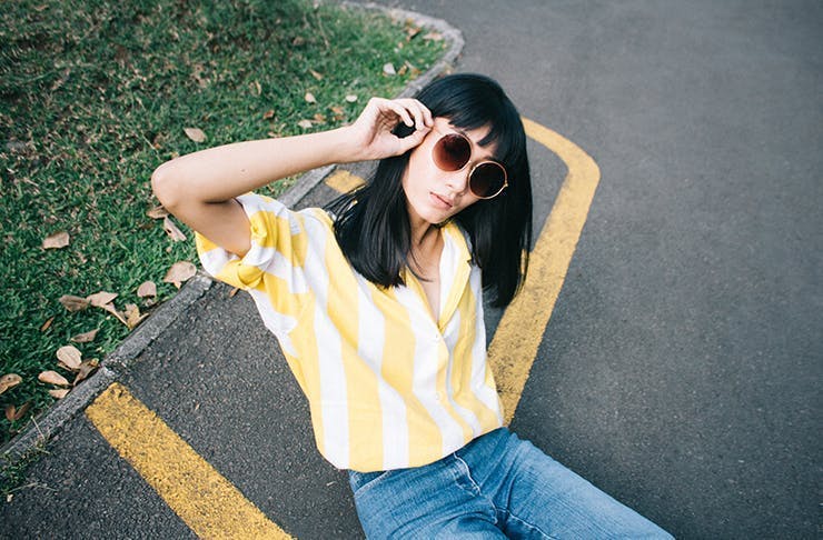 A woman wearing blue jeans and a yellow top sits on the pavement. She has a dark hair with a fringe and is wearing sunglasses. 