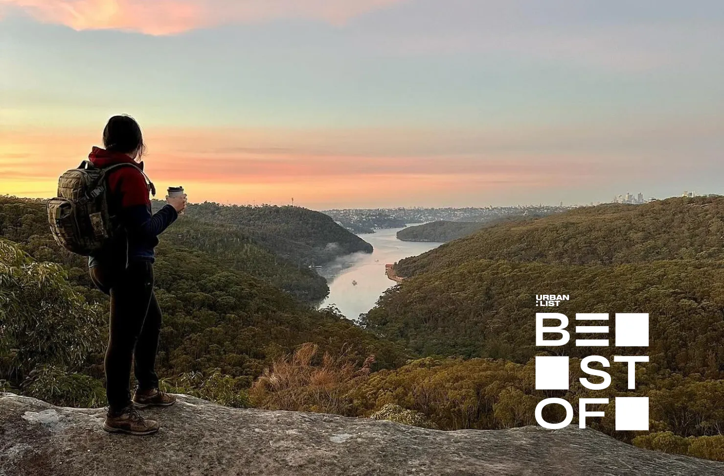 A hiker wearing a backpack stands on a rocky lookout at sunrise, holding a takeaway coffee while overlooking a forested valley, winding river and pastel sky.