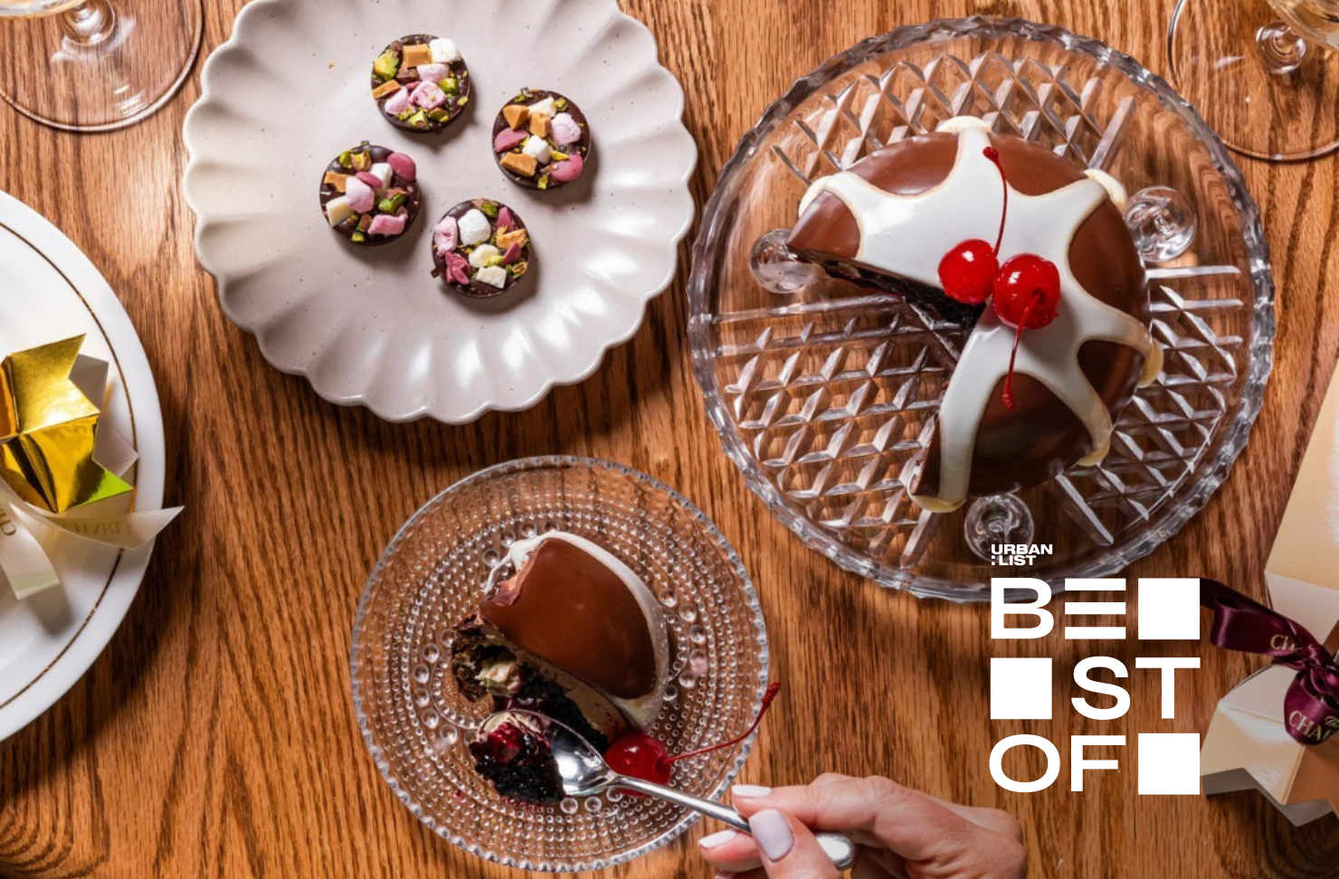 Overhead view of a festive dessert spread on a wooden table featuring a glossy chocolate Christmas pudding topped with cherries, bite-sized chocolates with nuts and marshmallows, and a hand holding a spoon ready to take a bite.