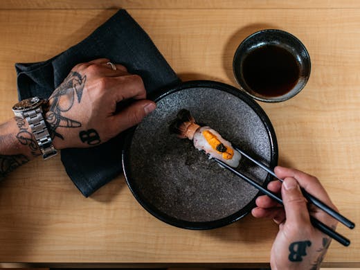 A person with tattoos picking up sushi with chopsticks 