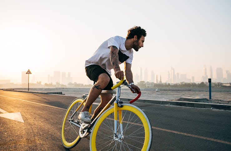 man in shorts and t-shirt riding a yellow and white road bike on a road, with a river and city skyline behind him in the distance