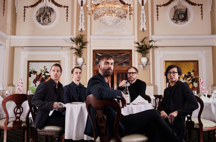 The band members of Birds of Tokyo sitting at seperate tables in a baroque style room.