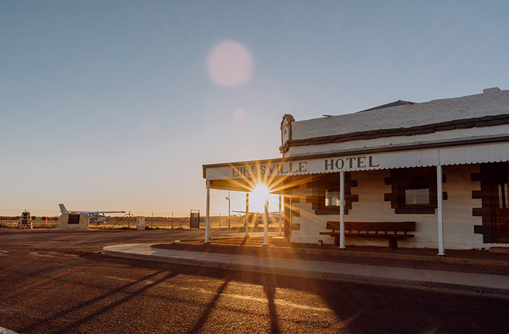 The Birdsville Hotel next to an airfield with light planes parked by a fence. The dirt is red and skies are clear.