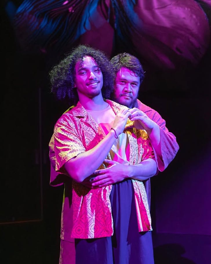 One man embraces another from behind under purple stage lighting.