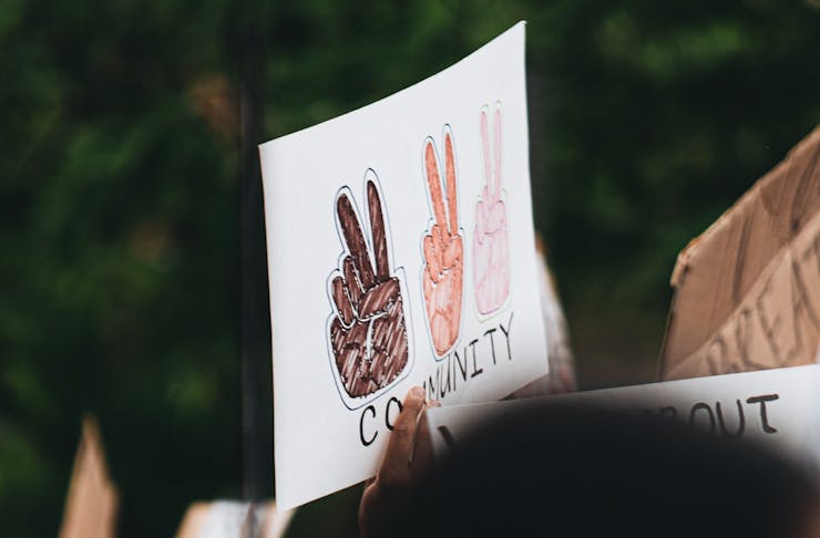 A sign being held up at a protest. The sign reads 'community' and has three different coloured hands doing a peace sign.