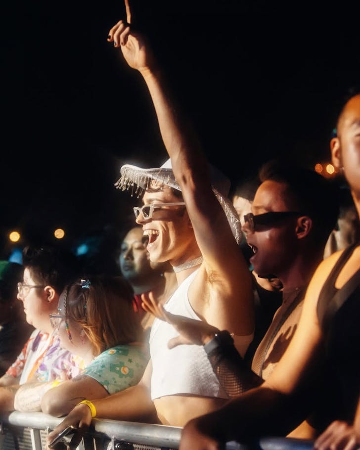 A lively crowd at a Mardi Gras event, with people dancing and cheering near the stage. One person in the foreground wears a white beaded hat, sunglasses, and a white crop top, raising an arm in excitement under bright stage lights.