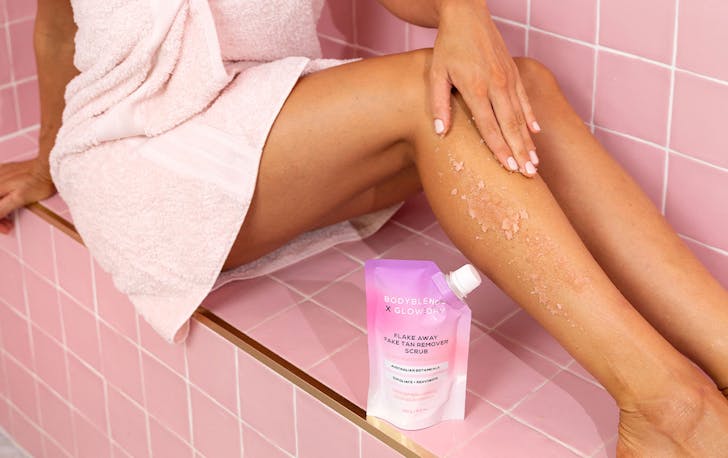 a woman sitting on a bathroom bench rubbing exfoliator onto her legs