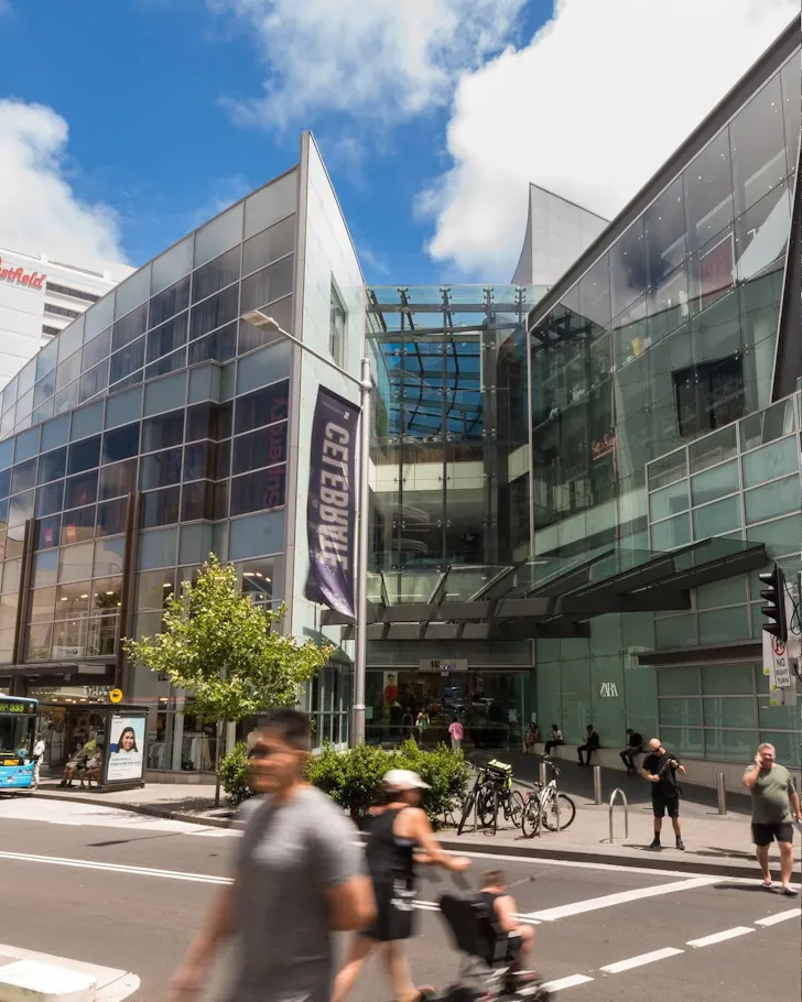 Pedestrians walk past the glass-fronted entrance to Bondi Junction's retail precinct, surrounded by shops, cafés and modern commercial buildings.