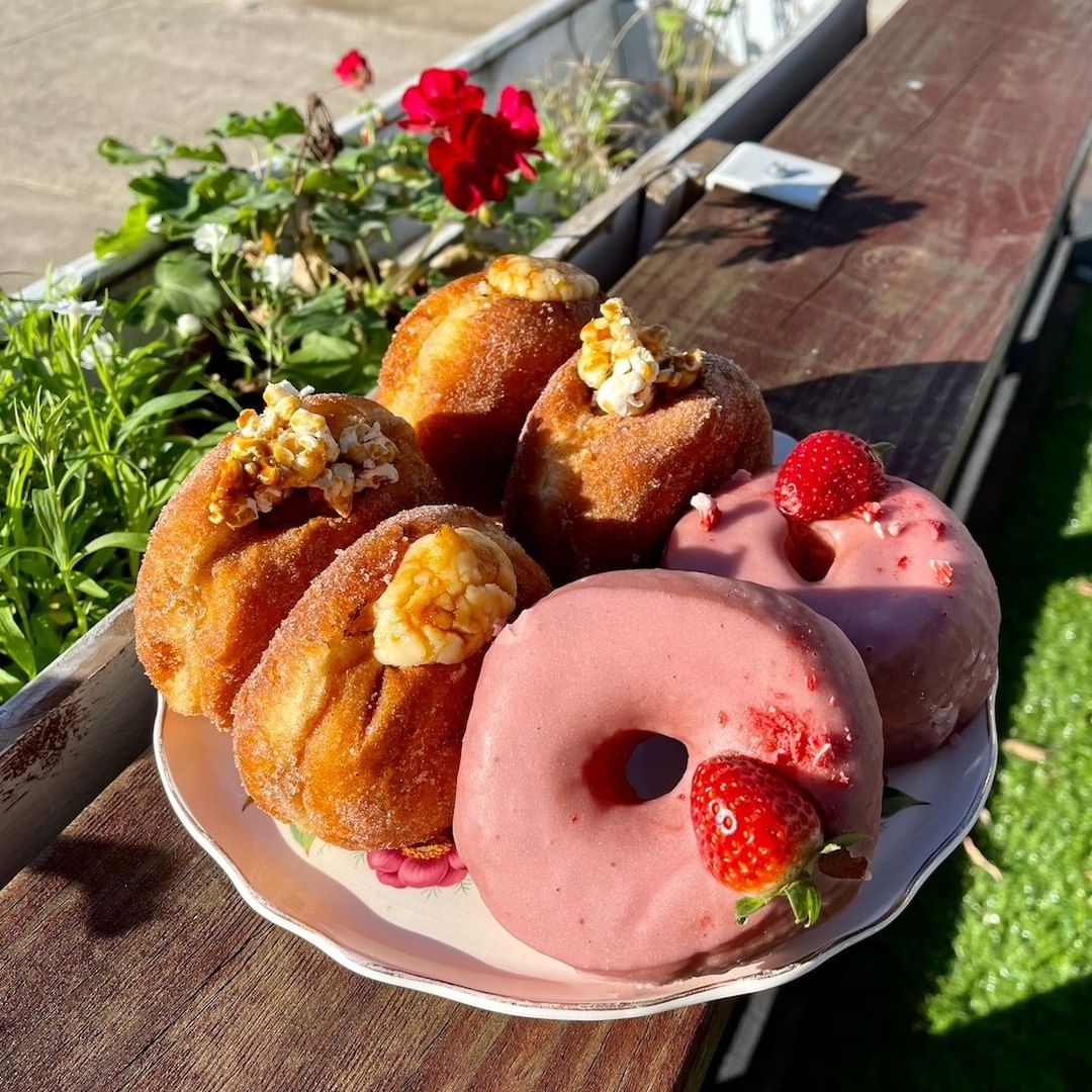 Six donut sitting on a plate outside. Two are glazed with pink icing and strawberries