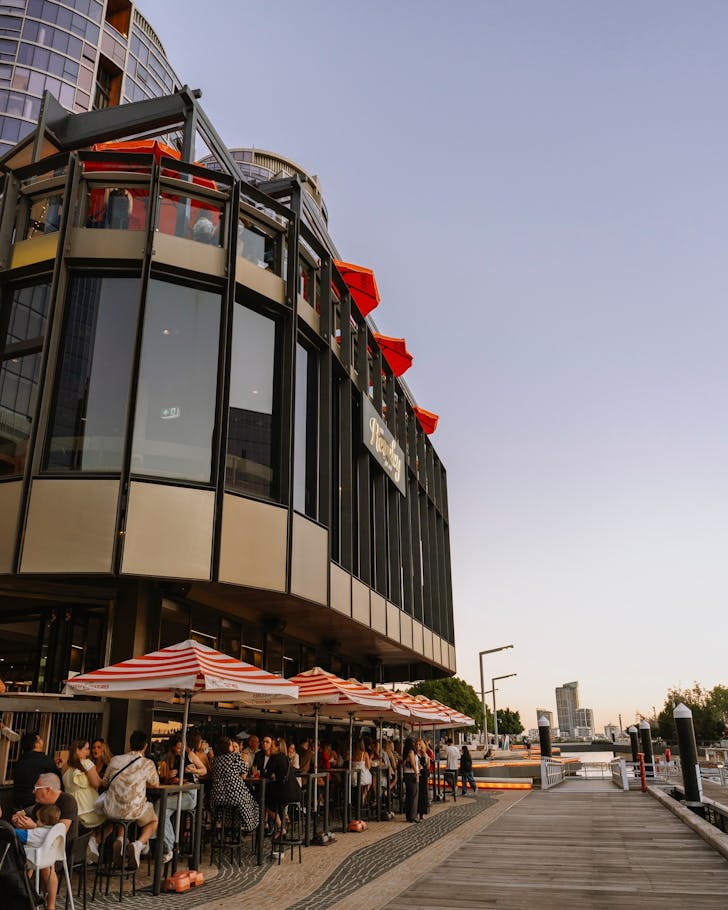 âRiverside dining at The Reveley in Elizabeth Quay Perth, with people enjoying drinks under red-and-white umbrellas at sunset.â