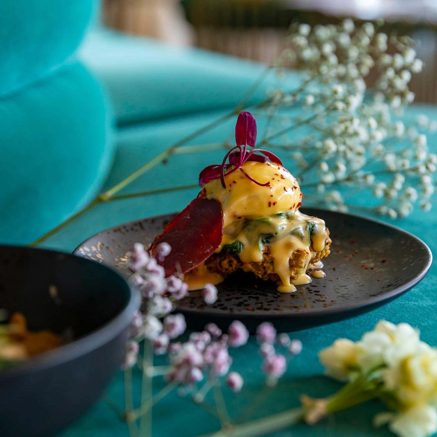 A close-up shot of a colourful brunch egg-dish, garnished with purple flowers and dripping with Hollandaise sauce.