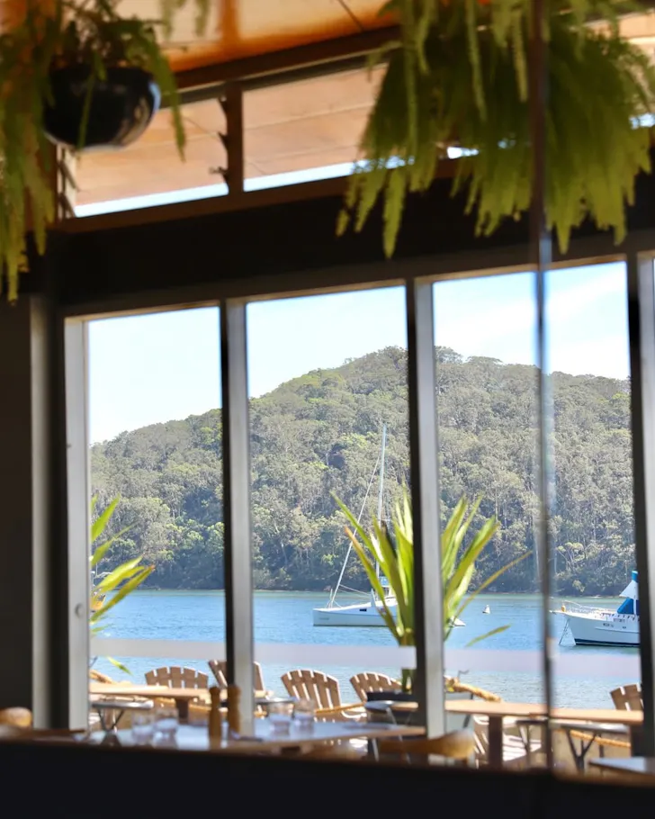 Dining room with floor-to-ceiling windows overlooking a calm bay with sailboats, outdoor seating and lush bushland on the Central Coast.