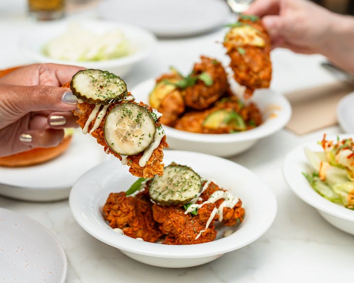Someone pulls a piece of cucumber-topped fried chicken from a bowl, amongst a table of fried chicken dishes.