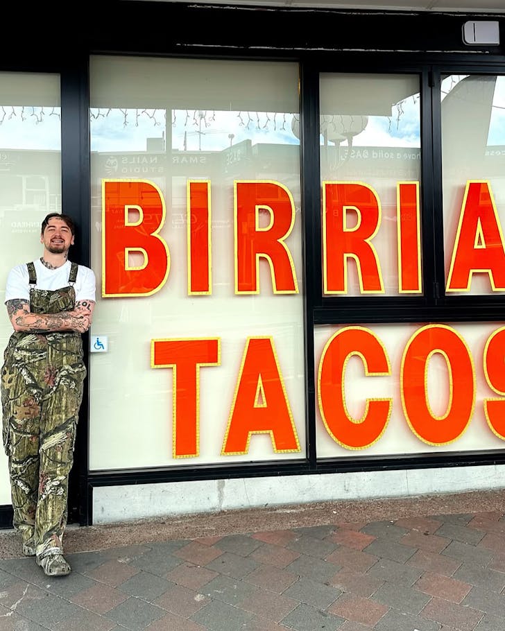 Sean Yarbrough wears camouflage overalls and leans up against a the shopfront of his second Broke Boy Taco store. 