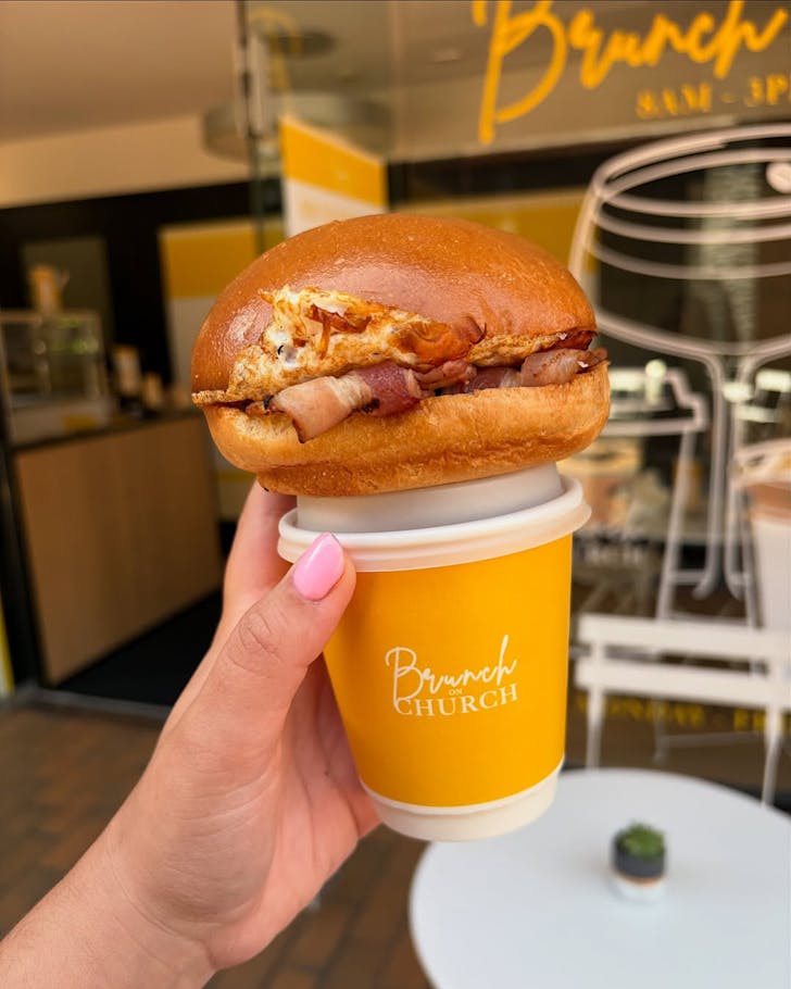 A bacon and egg brekkie bun sits on top of a bright yellow coffee cup. 