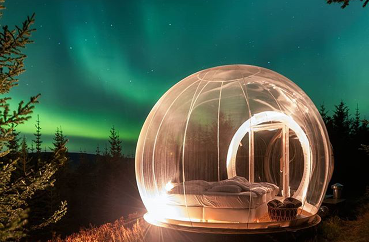 A clear bubble with a bed inside underneath the Northern Lights.