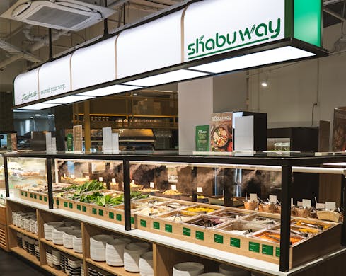 Inside Shabuway restaurant, a display window showcases the self-serve buffet food options. There is a large, lit-up sign hanging from the ceiling that has the Shabuway name and logo on it.